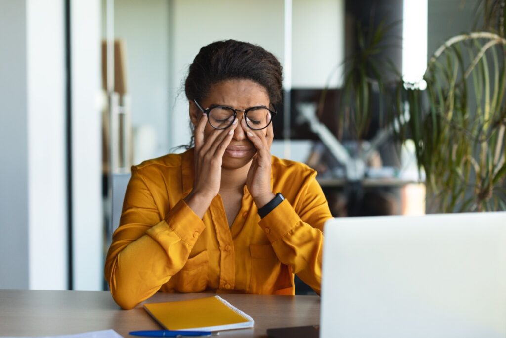 Woman wearing orange sweater sitting at her computer desk and rubbing her eyes under her glasses.