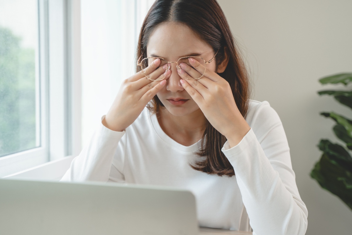 Woman wearing white shirt sitting at her computer desk and rubbing her eyes under her glasses.